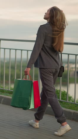 dama de gris caminando a lo largo del ferrocarril, sosteniendo bolsas de compras y bolso de mano negro, ajustando el cabello mientras camina con el fondo con vista borrosa del puente lejano sobre el río