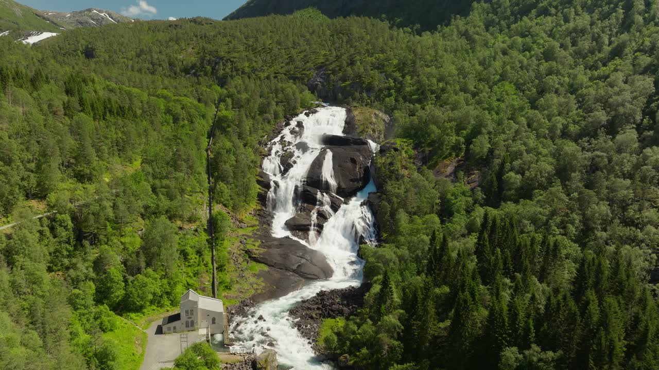Dron zachycuje mohutný vodopád Tveitafossen v norském údolí Husedalen, zdůrazňující vodní elektrárnu obklopenou dramatickými horami a scenérií tekoucí řeky.