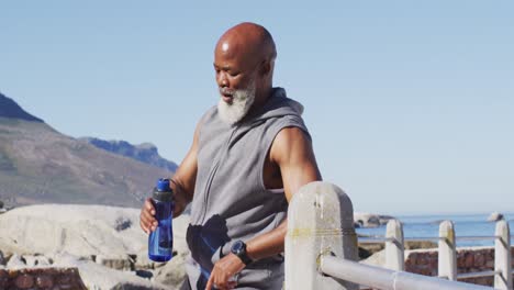 Senior-african-american-man-exercising-drinking-water-on-rocks-by-the-sea