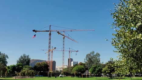 grúas para la construcción en la ciudad vistas después de un parque, árboles verdes bordean el marco filmado en cámara lenta de 4k