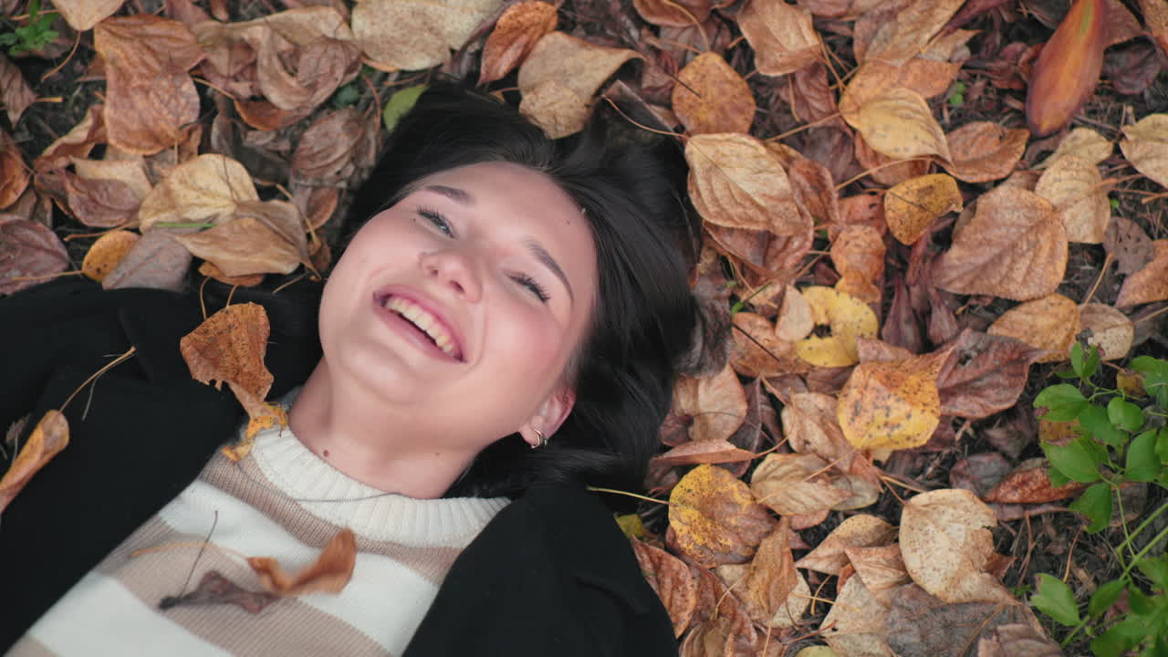 Overhead view of relaxed woman lying on forest floor laughing out loud while dry leaves fall around her, cozy sweater and black coat, carefree autumn mood captured with soft natural light