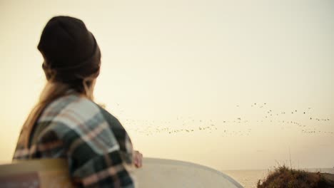 Vista-Trasera-De-Una-Niña-Rubia-Con-Un-Sombrero-Verde-Y-Una-Camisa-A-Cuadros-Parada-Cerca-Del-Mar-Con-Una-Tabla-De-Surf-En-Sus-Manos-Y-Mirando-Una-Gran-Bandada-De-Pájaros-Que-Vuelan-En-Algún-Lugar-En-El-Otoño.