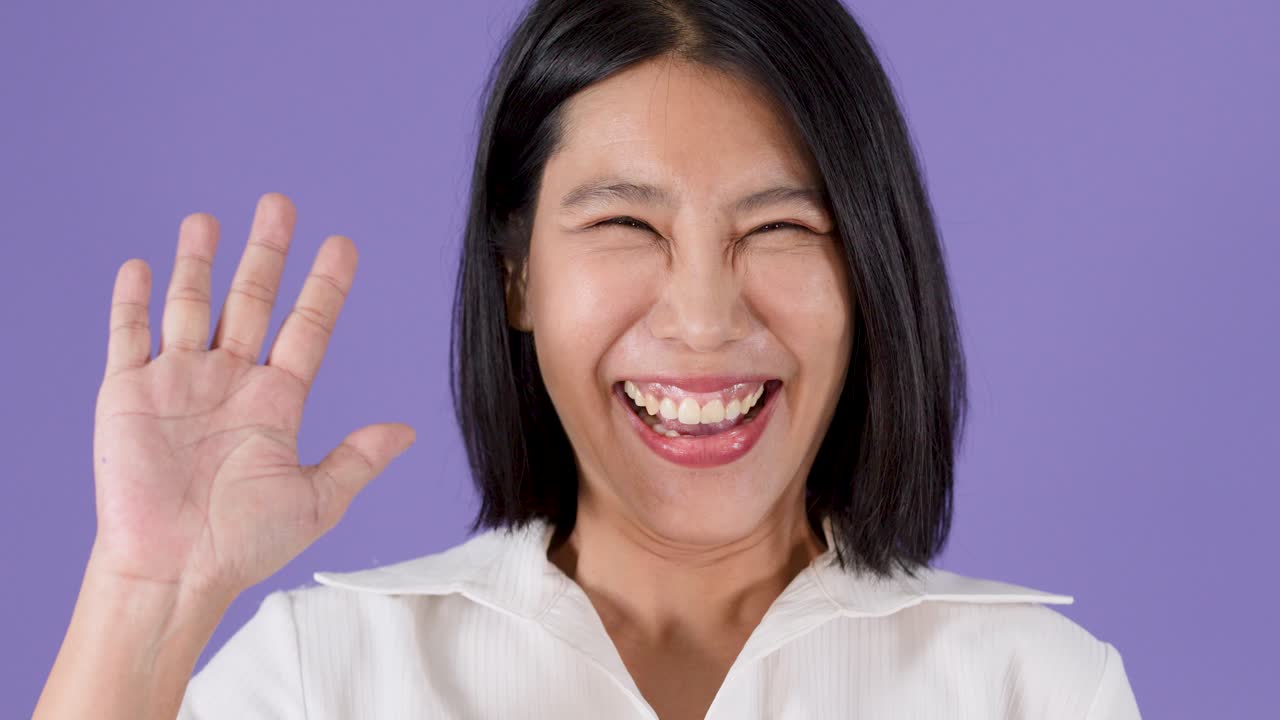 보라색 스튜디오 배경에서 짧은 검은 머리의 발랄한 아시아 여성이 미소를 지으며 손을 흔들고 있어요.