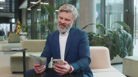 successful businessman holding smartphone and counting dollar bill money fan sitting in office lobby