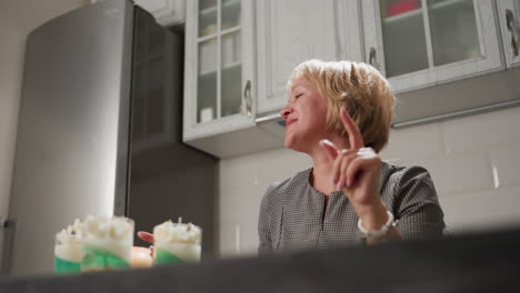 cheerful woman sings and dances joyfully in bright kitchen, moving hands expressively beside tall freezer and white painted cabinets, celebrating happy moment with delicious homemade desserts