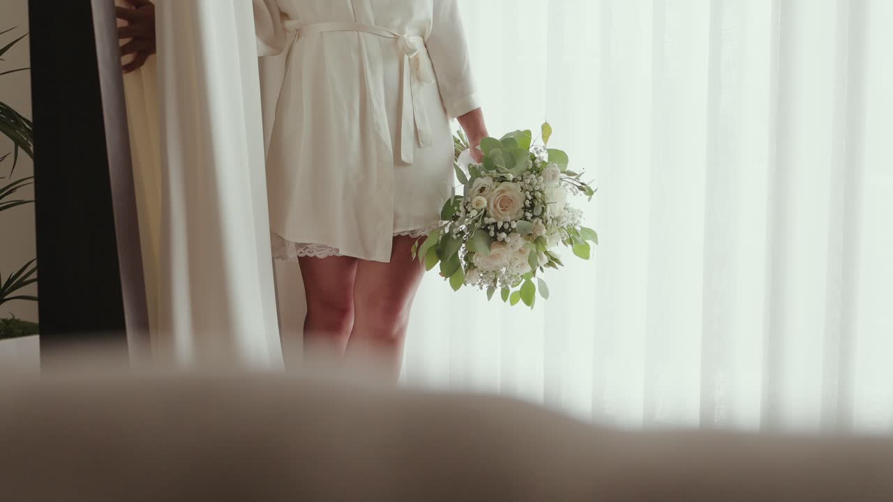 Close up of a woman in a robe holding a floral bouquet near bright white curtains
