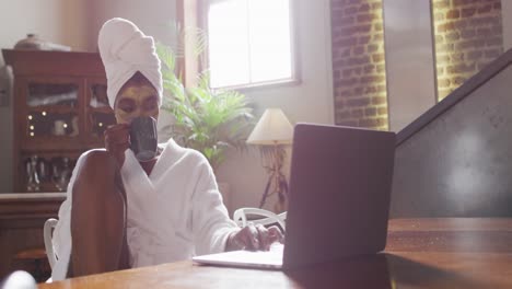 mujer atractiva afroamericana con máscara facial aplicada bebiendo café y usando computadora portátil en casa