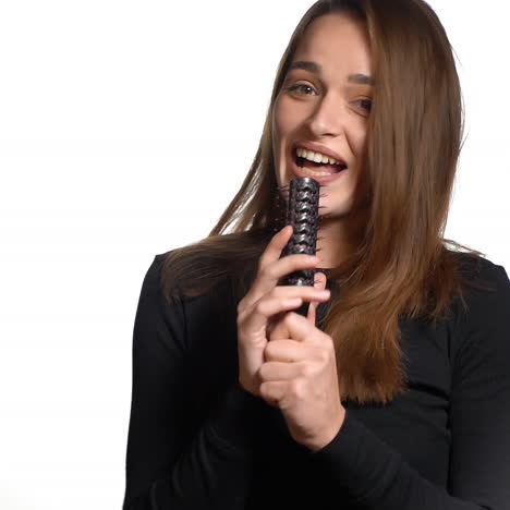 a joyful girl in a black sweater is combing her hair and starting to sing a song, holding a hairbrush as a microphone on a white background in the studio.