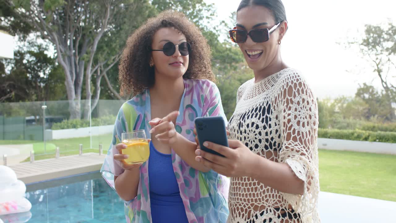 African American women standing by pool holding orange drinks while right woman sharing phone post
