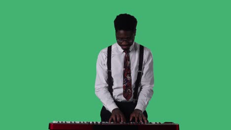 portrait of young african american man smiling and playing slow melody on a keyboard synthesizer. black male with tie and glasses in white shirt posing on green screen in studio. close up