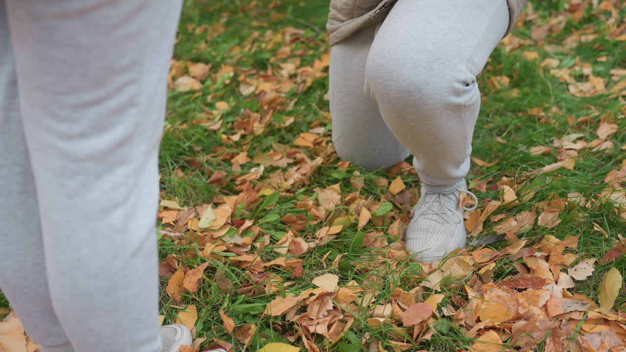 Lower body view of people wearing same outfit during proposal moment with one person kneeling on grass surrounded by dry autumn leaves and green foliage in peaceful outdoor setting