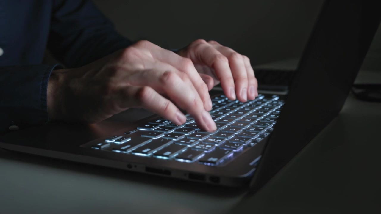 Dita pronte a digitare, mani che compongono un'email alla scrivania in una stanza in penombra, tastiera retroilluminata e polsino della camicia
