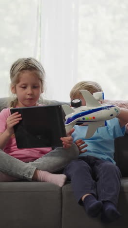 toddler boy plays with airplane toy while preschooler girl watches educational videos on tablet. siblings enjoy activities at home on weekend