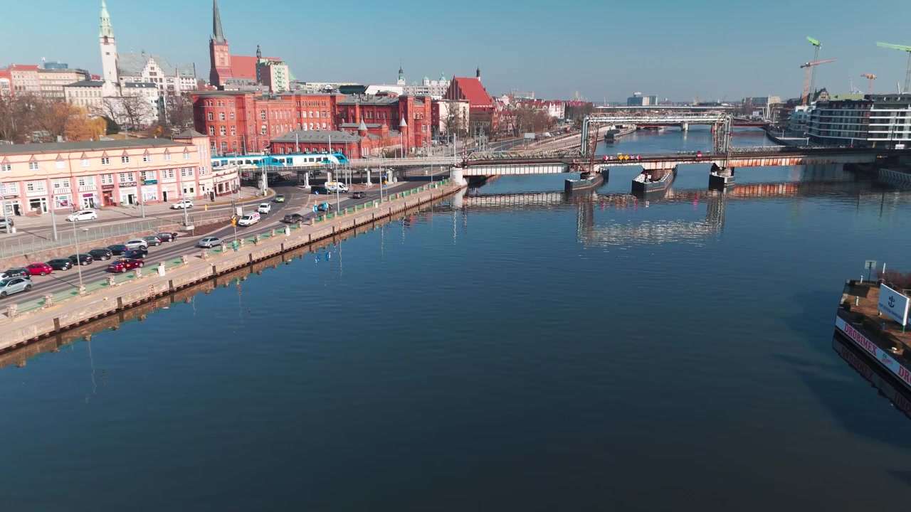 Una ripresa dall'alto che mostra un treno passeggeri che attraversa un ponte sul fiume Odra a Stettino, con l'architettura della città e una piccola barca in primo piano