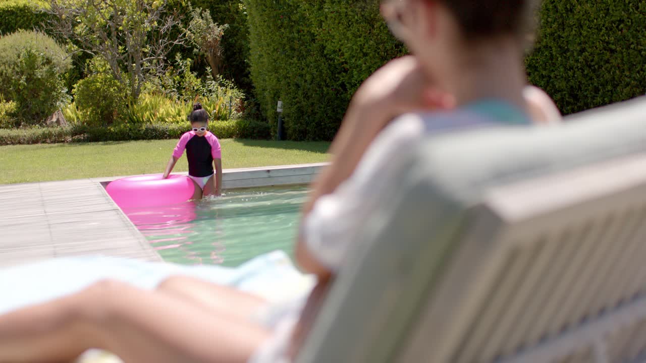 African American woman on chair watching female child easing pink tube, entering pool with goggles