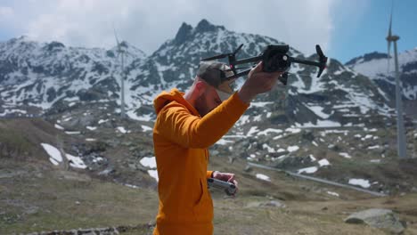 man with orange hoodie and cap holds drone while starting propellers and makes it fly with swiss mountains in background