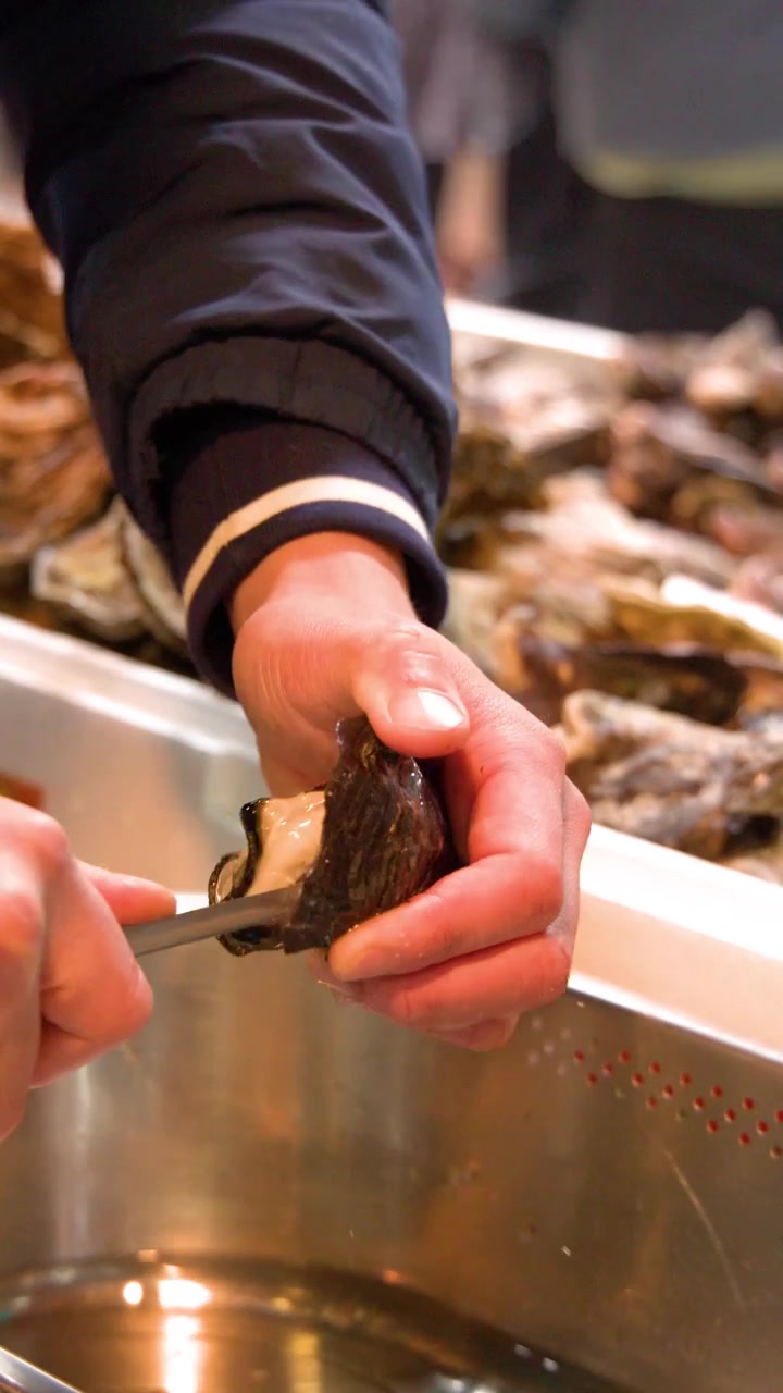 A vendor uses a knife to open fresh oysters at a busy outdoor fish market.