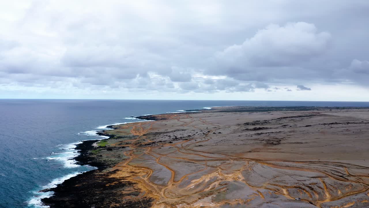 Expansive volcanic coastline with rugged dirt roads leading to the ocean. Waves crash against the rocky shore under an overcast sky, creating a remote, wild atmosphere.