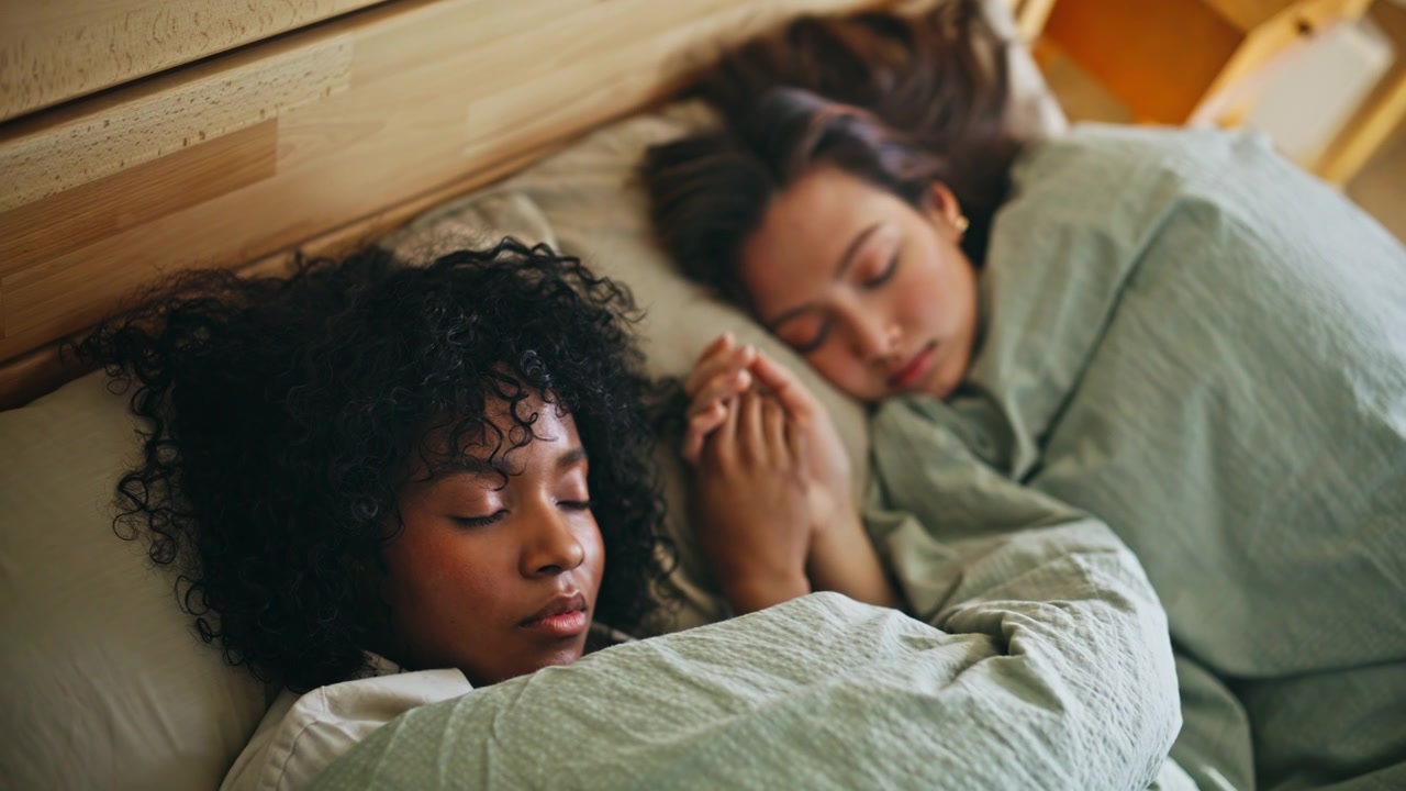 HBTQ-par sover hand i hand i en mysig säng. Kvinnor vilar hemma i sovrummet på morgonen