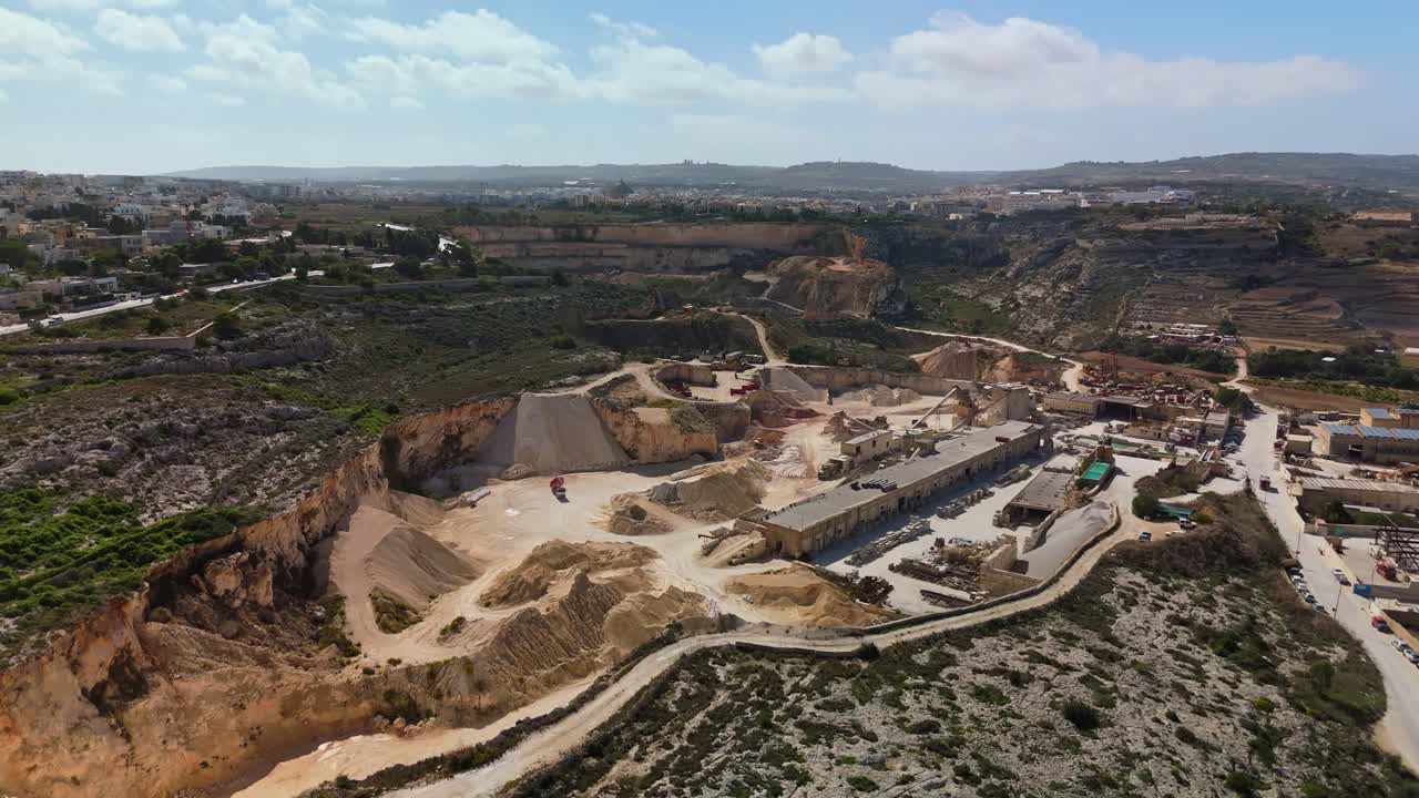Drone view showing a large active limestone quarry near Naxxar, Malta, with excavated rock formations, industrial machinery, and surrounding terraced countryside under a bright blue sky