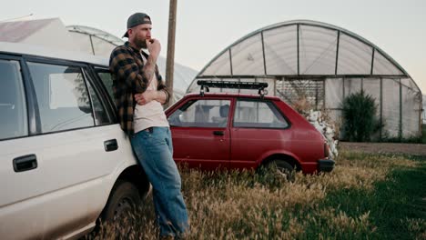 confident farmer guy in a cap leans on a white car and tries the tomato harvest on a farm among greenhouses