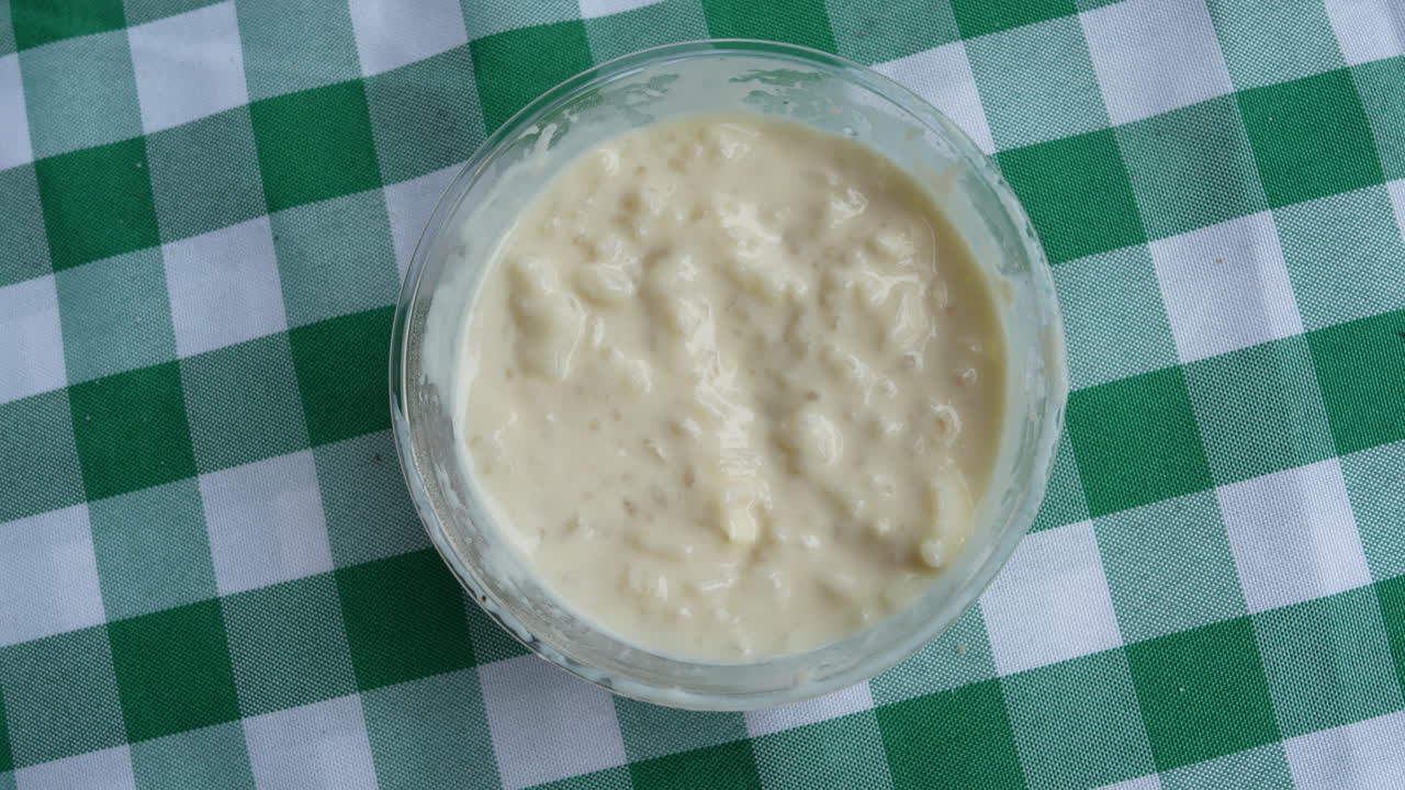 Delicioso arroz con leche colombiano, servido en un cuenco transparente sobre un mantel de cuadros verdes. El cremoso arroz con leche presenta una textura rica bajo luz natural, perfecto para temas culinarios.