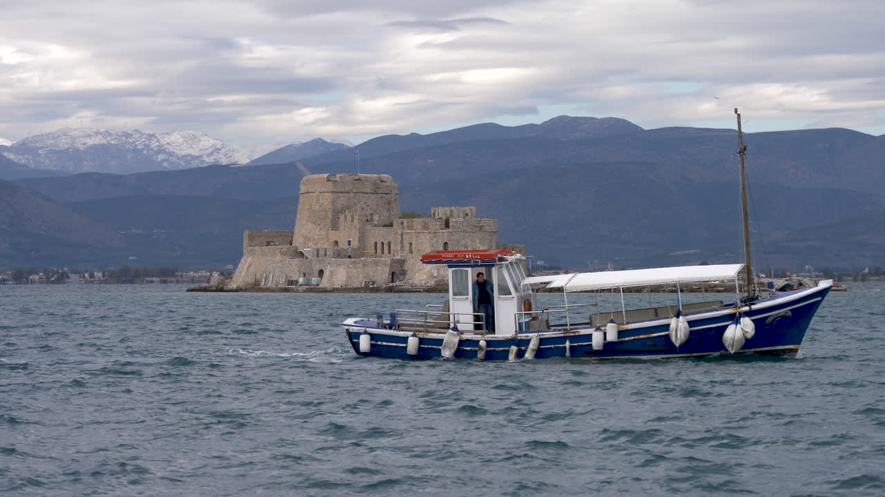 A blue fishing boat silently enters the port of Nafplio on Greek Peloponnese. In the background you can see the fort in the bay. Summer day.
