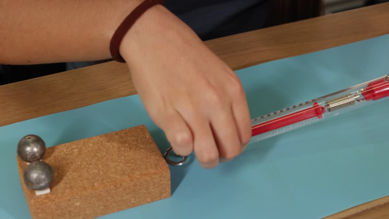 Hands use a spring scale to measure force on a weighted block, illustrating friction concepts on a smooth surface