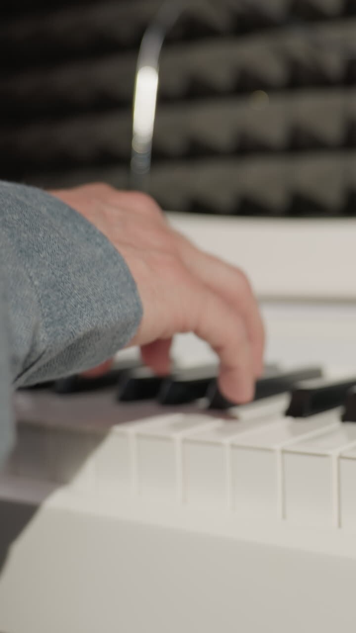 Close-up shot of a hand wearing a blue sleeve playing a piano, with a detailed focus on the fingers and keys. The background features an acoustic wall