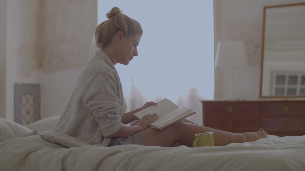 Focused woman sitting on bed and reading book after waking up