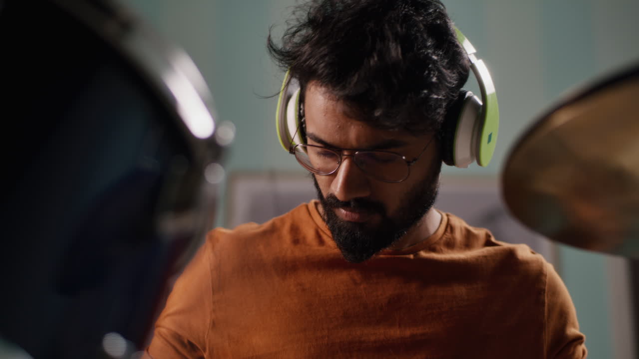 Indian Drummer Rehearsing at Home Bearded Indian Man in Glasses Listening to Music in Wireless Headphones and Playing Drums during Rehearsal in Home Studio
