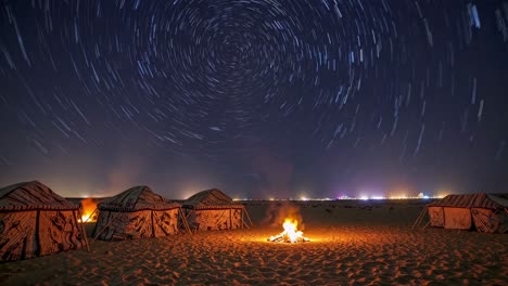 campfire glowing softly near camping tents, illuminating sandy landscape with mesmerizing star trails swirling overhead against pitch black nocturnal wilderness background
