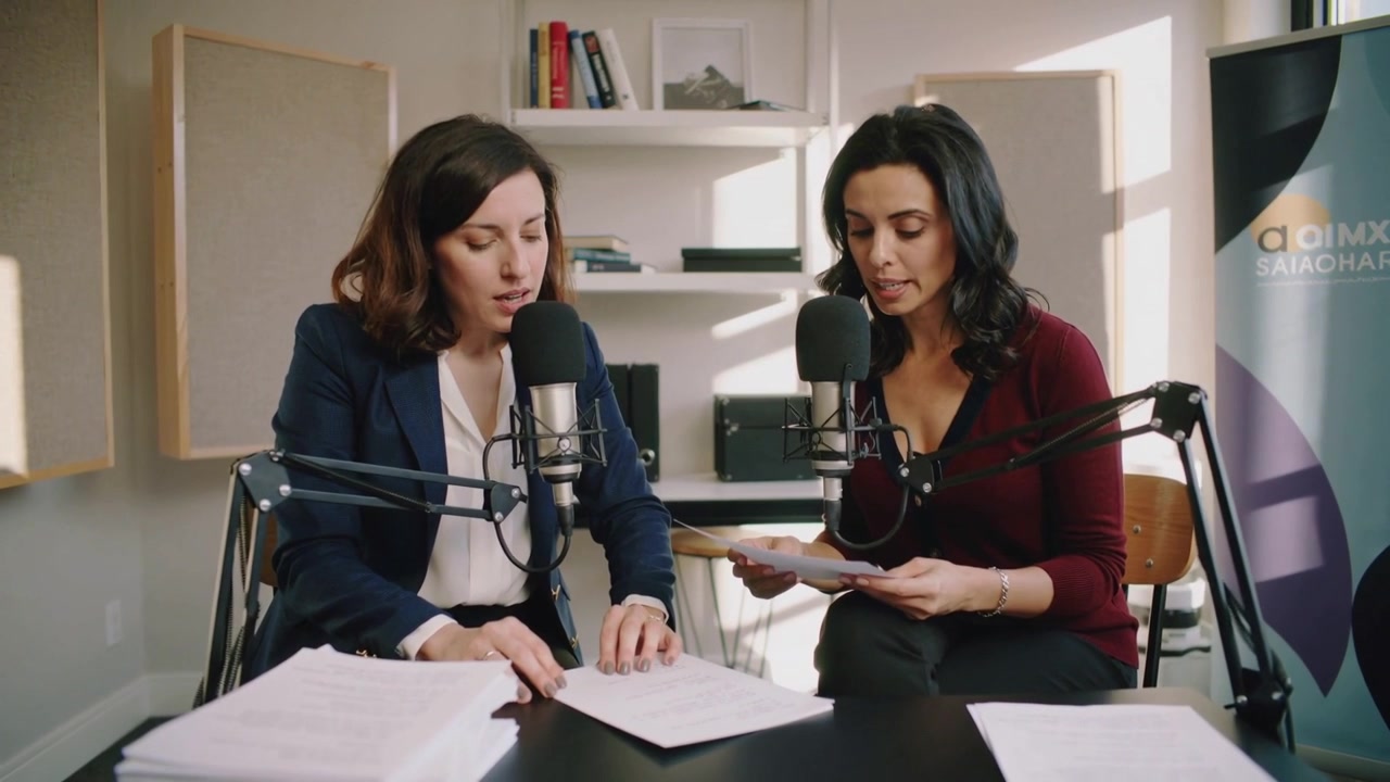 In studio per un podcast, il conduttore in blu scuro ordina una pila di copioni mentre il conduttore in bordeaux legge al microfono.