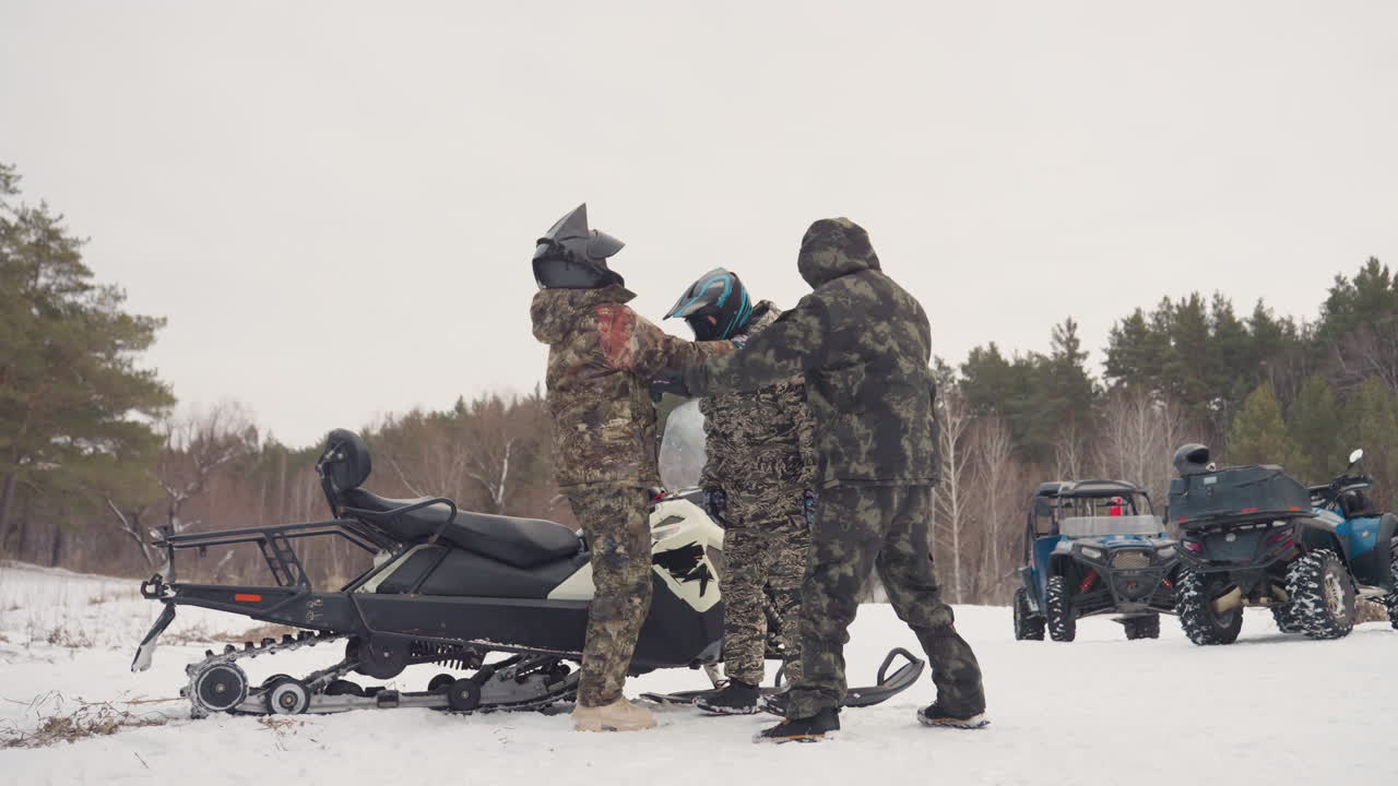 Un trio di piloti cammina accanto a un convoglio di motoslitte attraverso una radura ghiacciata, tute e caschi mimetici, il leader indica il sentiero mentre un pilota di supporto trasporta l'equipaggiamento, un orizzonte invernale drammatico e una linea di pini sullo sfondo.