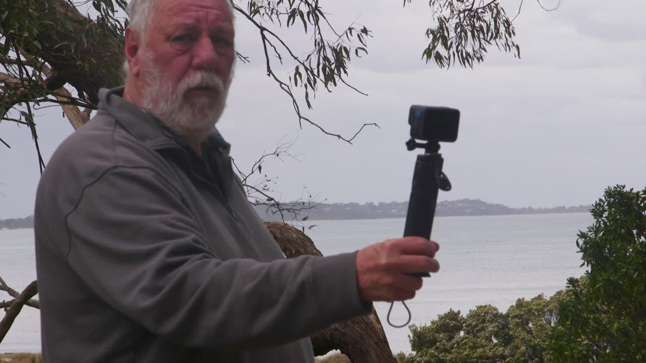 An elderly male digital creator captures beautiful natural scenery with his smartphone. The atmosphere is calm and peaceful, with a picturesque view of water and trees around him