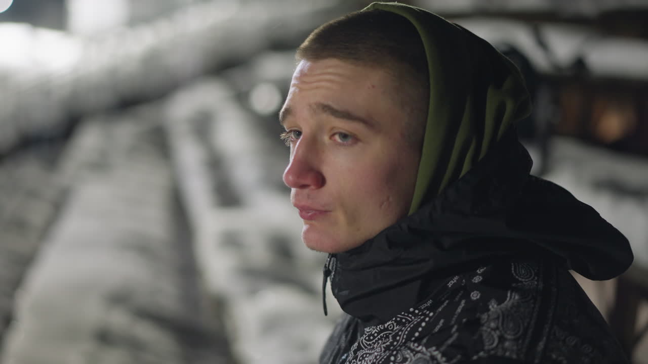 Side profile of street boy in hoodie looking around under night light near snowy seating area, with cold air visible on face and soft lighting casting reflective tones over winter surroundings