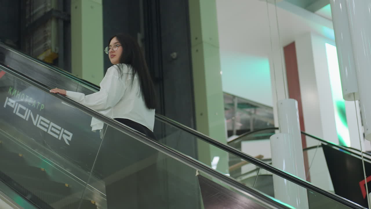Student wearing glasses and white shirt rides ascending escalator in modern mall, looking back with hand on glass railing, surrounded by bright lights, white columns, and clean architecture