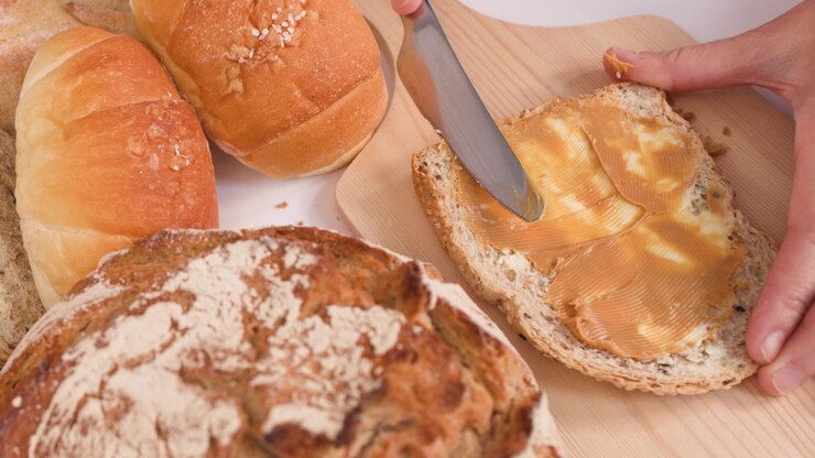 Close-up shot of spreading peanut butter on bread amidst sourdough, baguettes, and croissants.