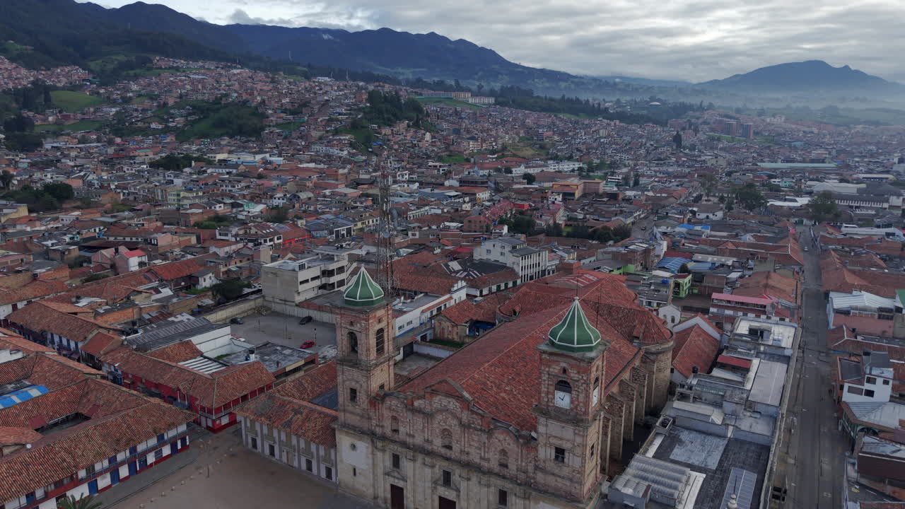 A breathtaking aerial view of Zipaquira's cityscape featuring the iconic cathedral amidst a sea of terracotta rooftops, with majestic mountains in the background under an overcast sky