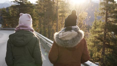 Two-female-tourists-walking-in-slow-motion-on-a-long-road-through-the-countryside-towards-the-snowy-mountain-peaks-and-golden-trees-on-the-background.-Rare-view