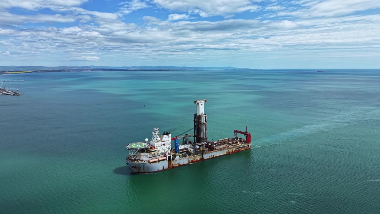 Industrial dredging vessel operates in open water near a coastal area with cloudy skies and distant land visible