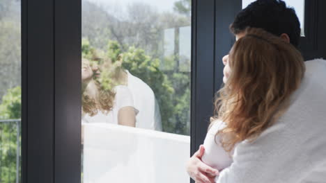 happy caucasian lesbian couple embracing, looking by window and smiling in sunny house, copy space