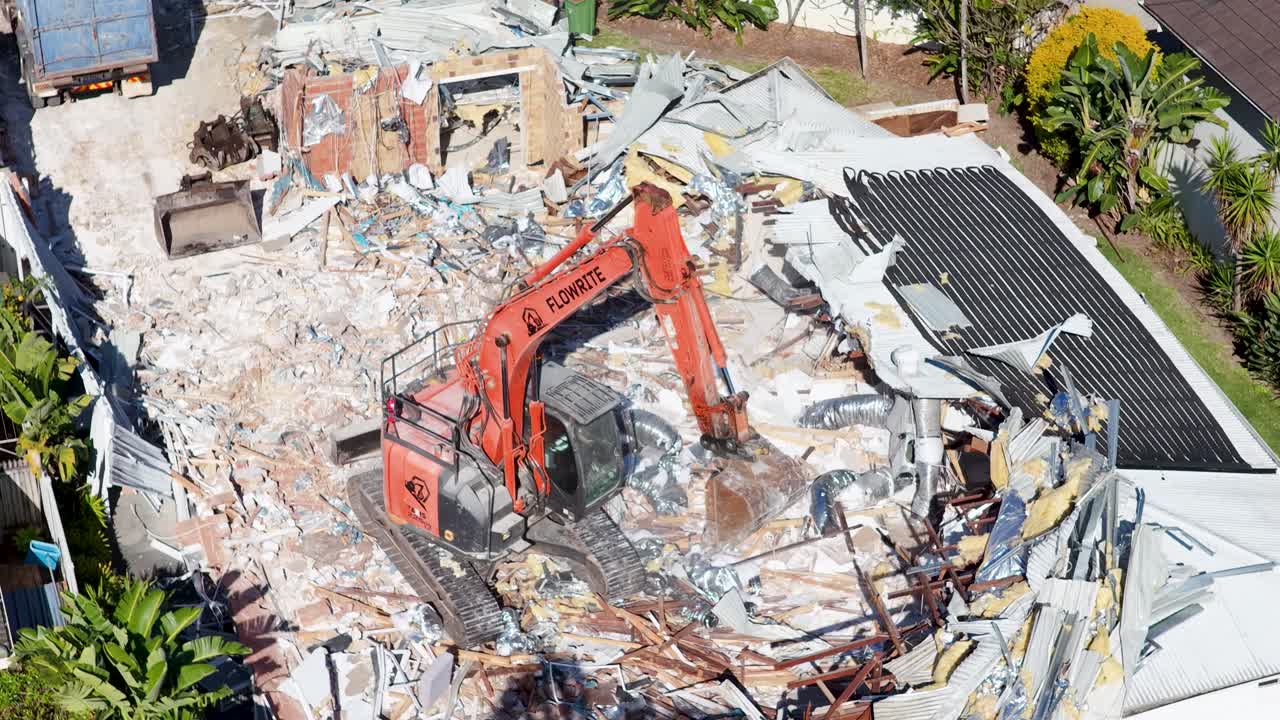 Aerial view of an orange excavator actively demolishing a house, surrounded by debris and vegetation, under bright daylight with steady drone camera movement