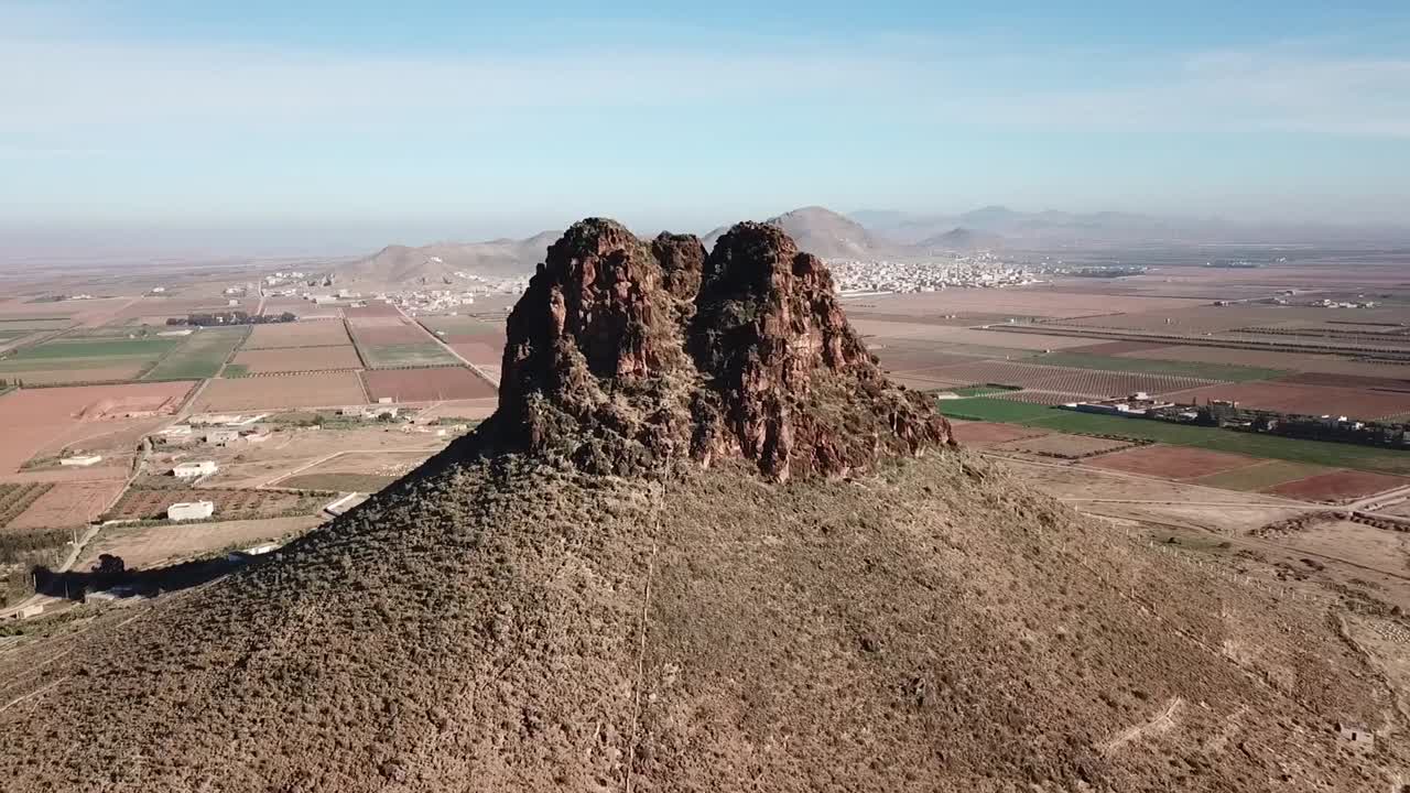 Cinematic drone shot performing a slow circular orbit around a unique rocky mountain peak standing atop a hill, overlooking vast agricultural plains and the countryside landscape