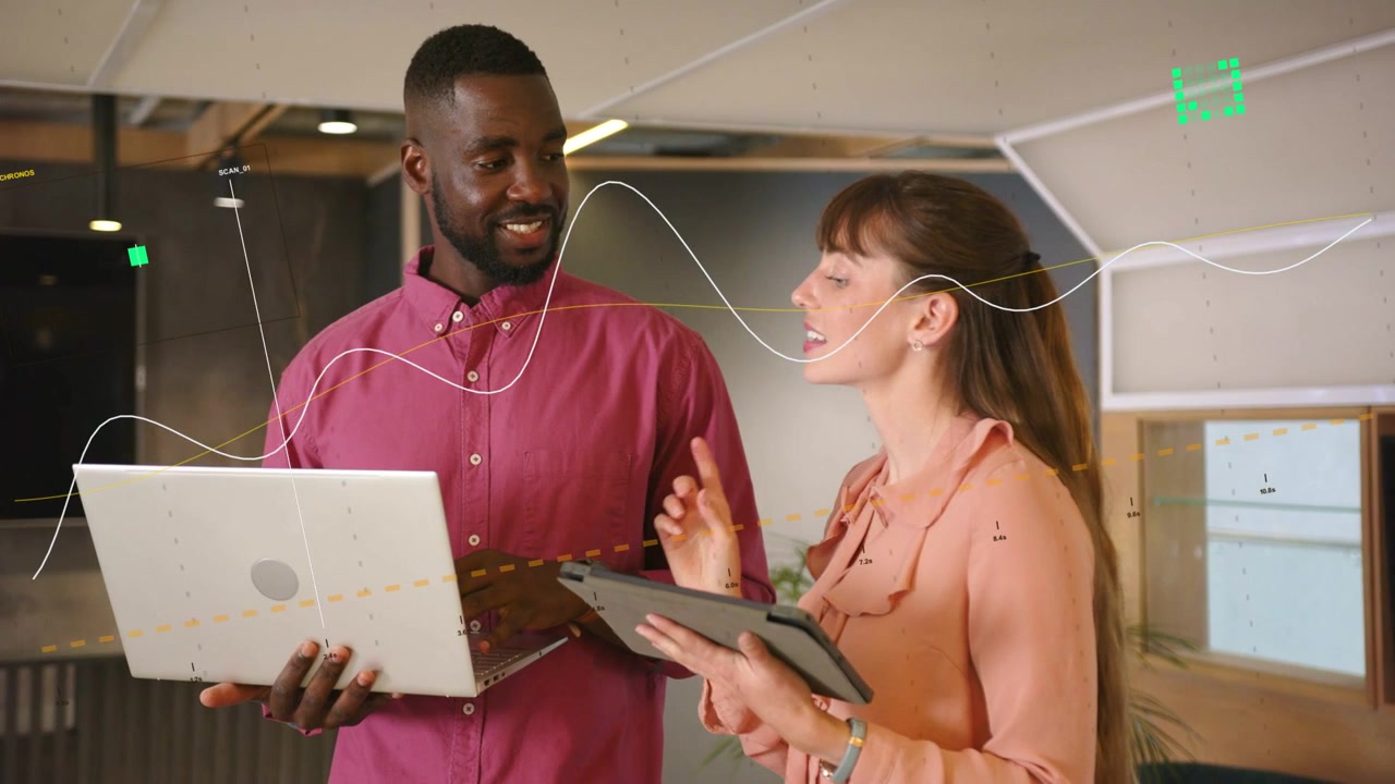 Woman speaking and pointing at tablet, man tapping laptop, reviewing business data with green icons