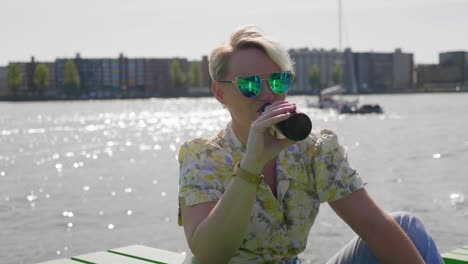 Casual-young-woman-drinks-a-beer-sitting-on-a-pier,-river-and-boats-in-the-background