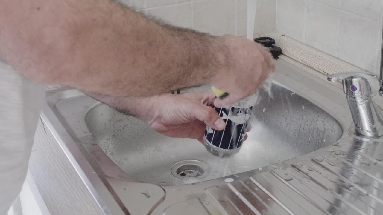 Man's hand washing a glass under running water in stainless steel kitchen sink. Close-up shot captures domestic cleaning task with natural light. Home hygiene and household chores concept