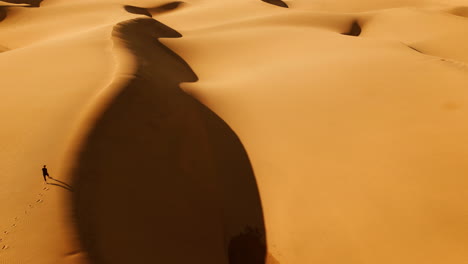 a stationary aerial view captures a man strolling along the ridge of the sand dunes, his figure contrasted against the vast desert landscape