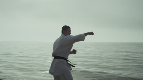 strong man practicing taekwondo near ocean against cloudy sky in slow motion.