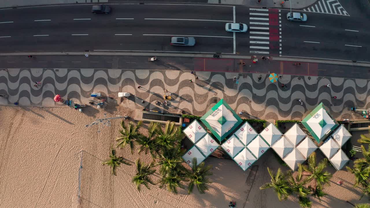 The Copacabana boardwalk buzzes with afternoon energy as elongated shadows from the surrounding mountains stretch across the iconic patterned promenade.
